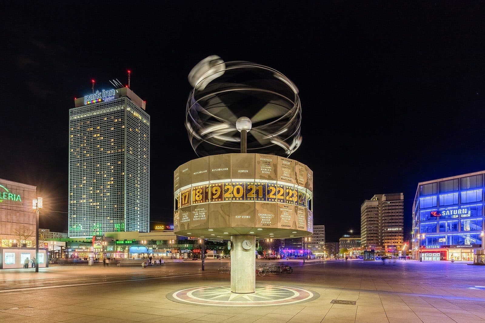 De Weltzeituhr op de Alexanderplatz bij avond, met verlichte gebouwen rondom