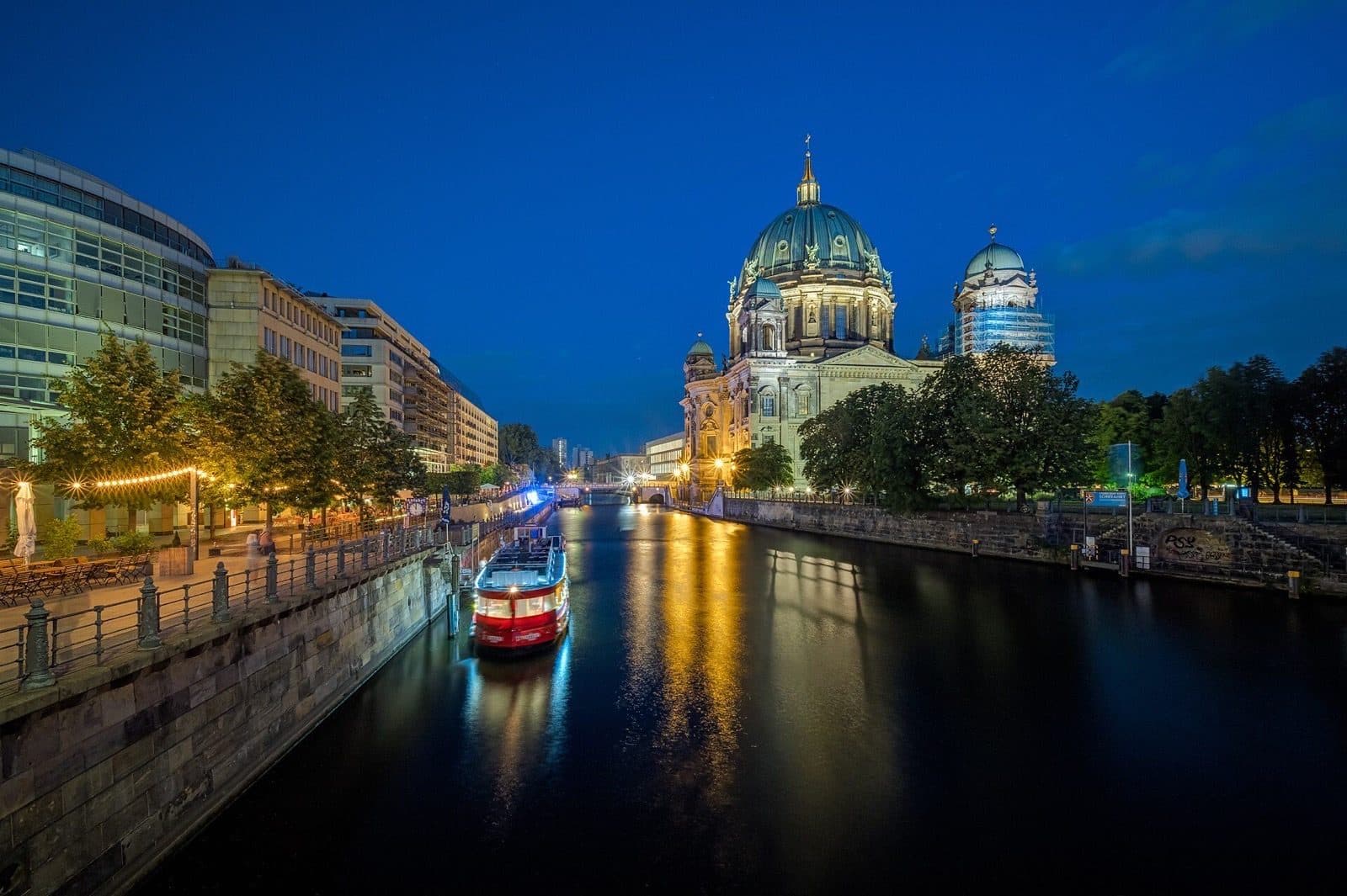 De verlichte Berliner Dom aan de Spree tijdens het blauwe uur, met een rondvaartboot op het water en terrasjes langs de kade