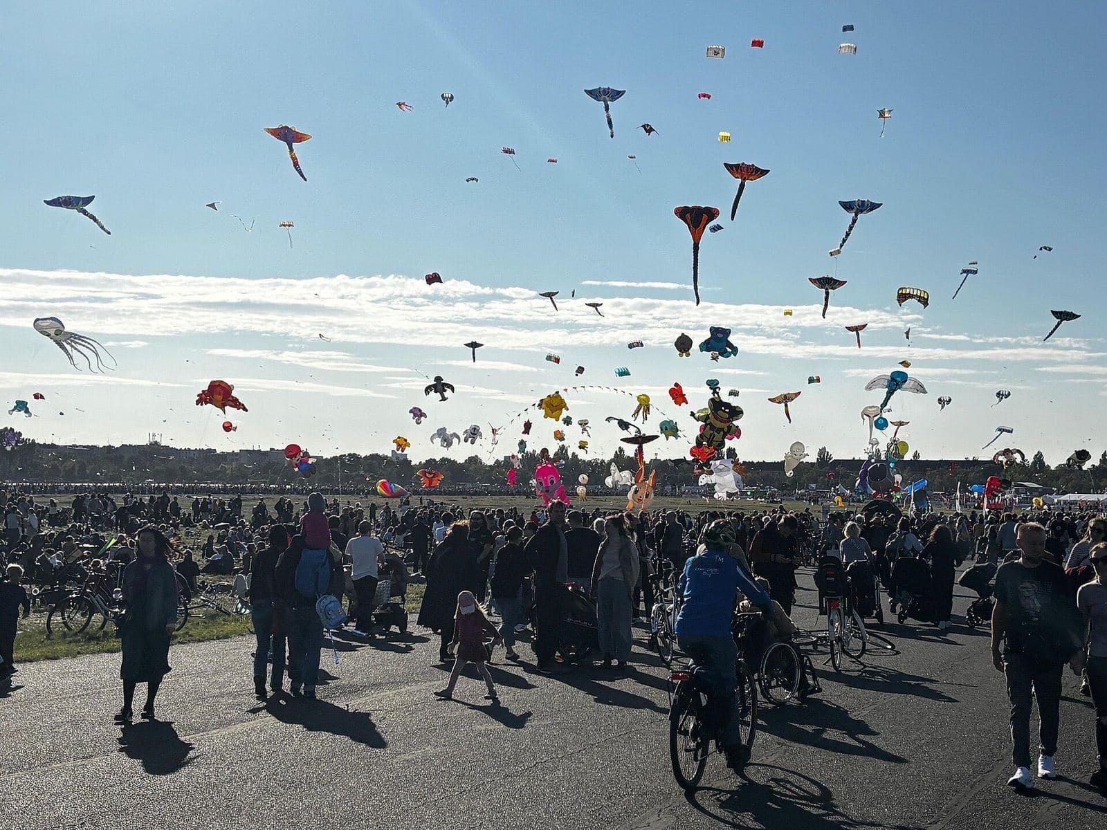 Honderden vliegers boven Tempelhofer Feld tijdens een zonnige middag, met mensen en fietsers op de baan